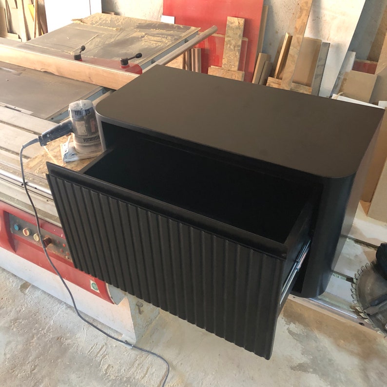 May include: Black wooden cabinet with a single drawer that has a ribbed front. The drawer is open and the cabinet is sitting on a workbench.