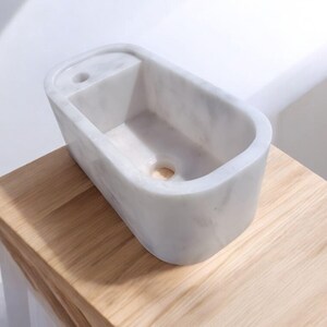 May include: A white marble sink with a rectangular basin and rounded edges. The sink has a small, rectangular shelf area on one side and a drain hole at the bottom. It is set on a light-colored wooden surface.