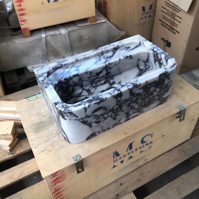 May include: A white and purple marble sink with a rectangular shape. The sink is sitting on a wooden crate with the text "MC MARBLESINK" printed on it.