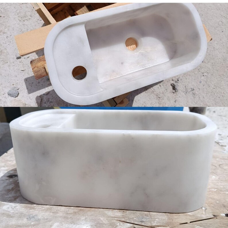 May include: A white marble sink with a rectangular basin and rounded edges. The sink has a built-in shelf and two circular drain holes. The smooth, polished surface of the sink reflects the light, highlighting the natural veining of the marble.