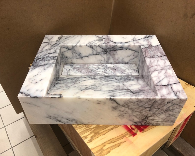 carrara-marble-wall-mount-sink-hero.jpg Finished rectangular Carrara White marble wall-mounted sink showing its polished white surface, soft gray veining and centered basin.