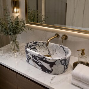 Puede incluir: Un lavabo ovalado de m&aacute;rmol blanco con vetas pronunciadas en color morado oscuro y negro. El lavabo est&aacute; montado sobre una encimera de m&aacute;rmol blanco con un grifo y manija de lat&oacute;n en la pared, junto a un dispensador de jab&oacute;n de vidrio y toallas.