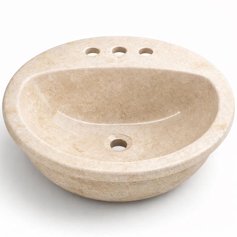 May include: A beige, oval-shaped bathroom sink made of stone. The sink has a rounded basin with three faucet holes and a single drain hole. The stone has a natural, textured appearance.