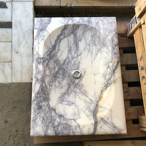Puede incluir: Un lavabo de m&aacute;rmol blanco con vetas moradas y un desag&uuml;e plateado.