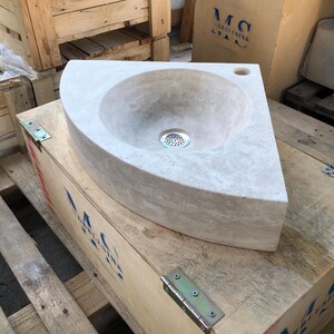 May include: A white corner sink made of stone with a round basin and a drain in the centre. The sink is sitting on a wooden crate.