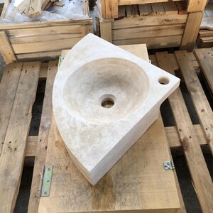 May include: A corner sink made of travertine stone. The sink is off-white with a smooth, polished finish. It has a rounded bowl and a single drain hole. The sink is sitting on a wooden pallet.