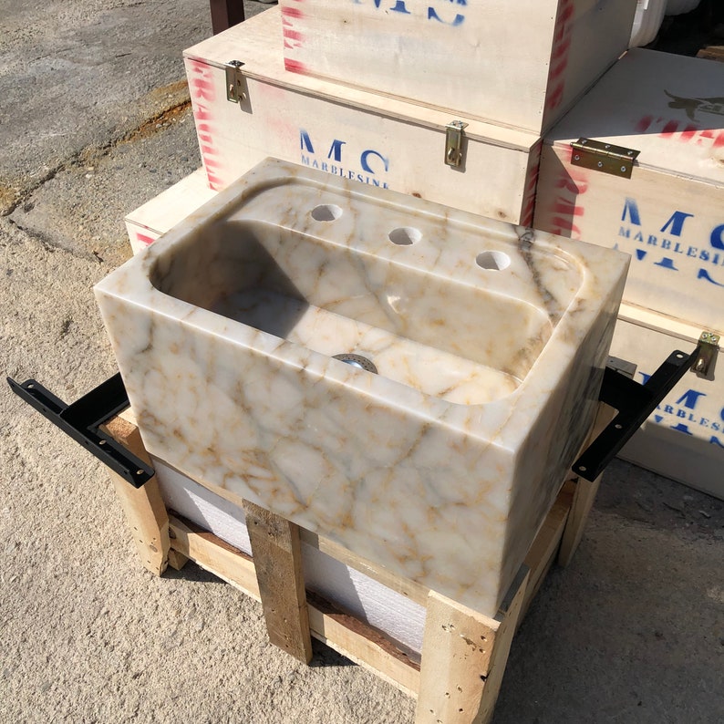 Front three-quarter view of a hand-carved Calacatta Viola marble bathroom sink with cream and gold veining, deep rectangular bowl, three faucet holes and black wall-mount brackets beside protective wooden crates.