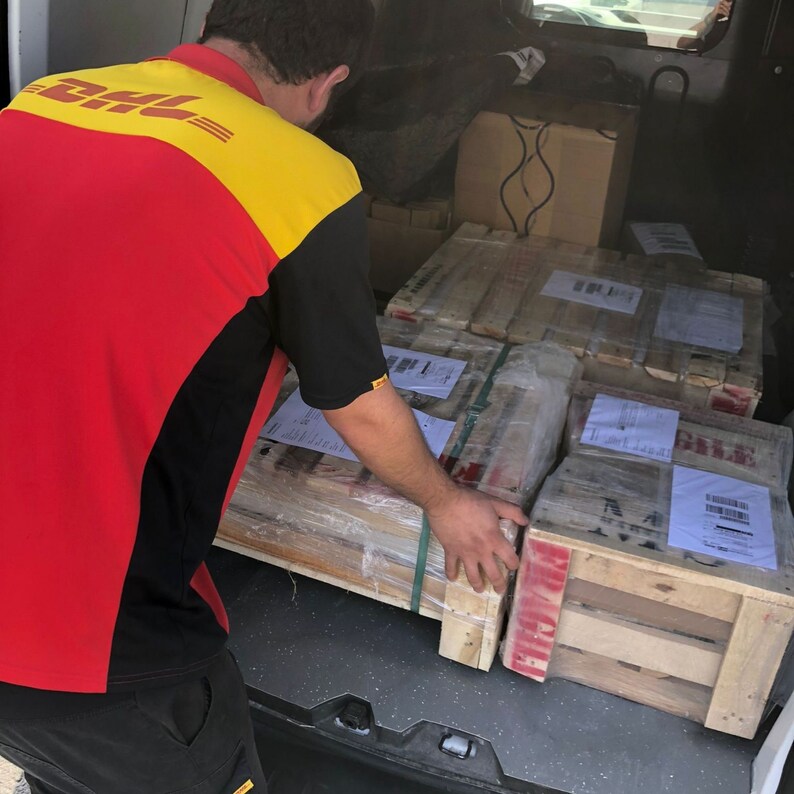 Może przedstawiać: Osoba w czerwono-ż&oacute;łtym uniformie DHL ładuje drewniane skrzynie do tylnej części samochodu dostawczego. Skrzynie są owinięte brązowym papierem i zabezpieczone taśmą.