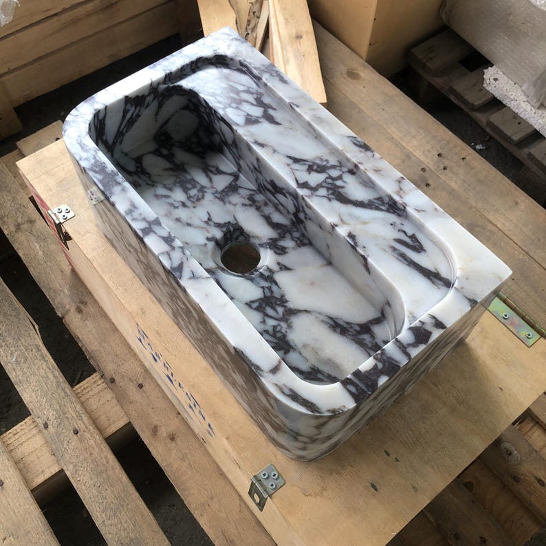 May include: A white marble sink with purple veining. The sink has two basins and a single drain hole. The sink is sitting on a wooden pallet.