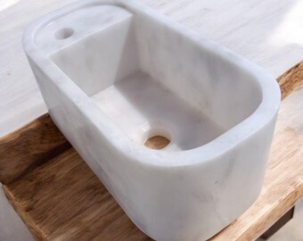May include: A rectangular white marble sink with rounded edges. The sink has a small rectangular section on the left side with a hole, and a drain hole at the bottom. The sink is placed on a wooden surface.