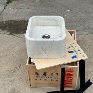 Side view of the Carrara White marble sink with black steel wall brackets, showing its compact rectangular form, thick stone construction and polished white surface.