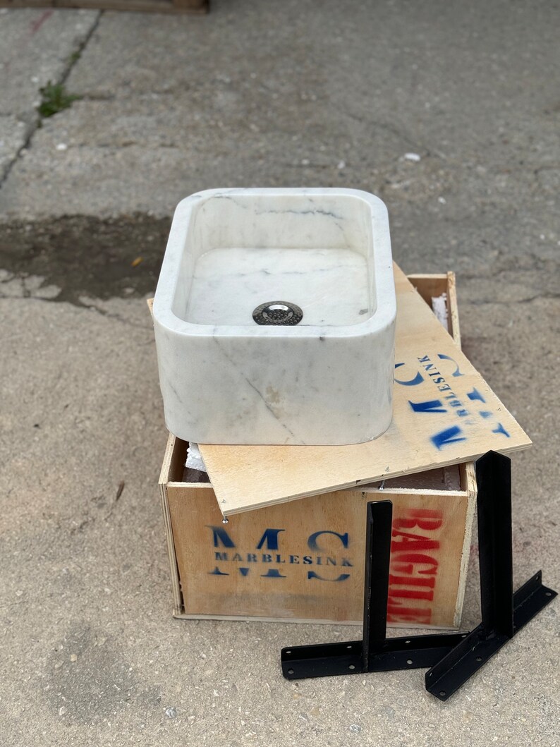 Side view of the Carrara White marble sink with black steel wall brackets, showing its compact rectangular form, thick stone construction and polished white surface.