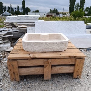 Op de afbeelding: Een rechthoekige beige stenen wastafel met een ruwe, getextureerde buitenkant en een gladde, gepolijste binnenkant. De wastafel heeft een enkel gecentreerd kraangat aan de achterrand en een natuurlijke, poreuze uitstraling van travertijn.