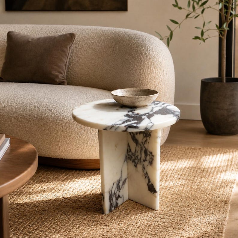 May include: A round side table made of white marble with prominent black veining, featuring a cylindrical top supported by a three-part intersecting base. A shallow, textured, beige ceramic bowl sits centered on the table's smooth surface.