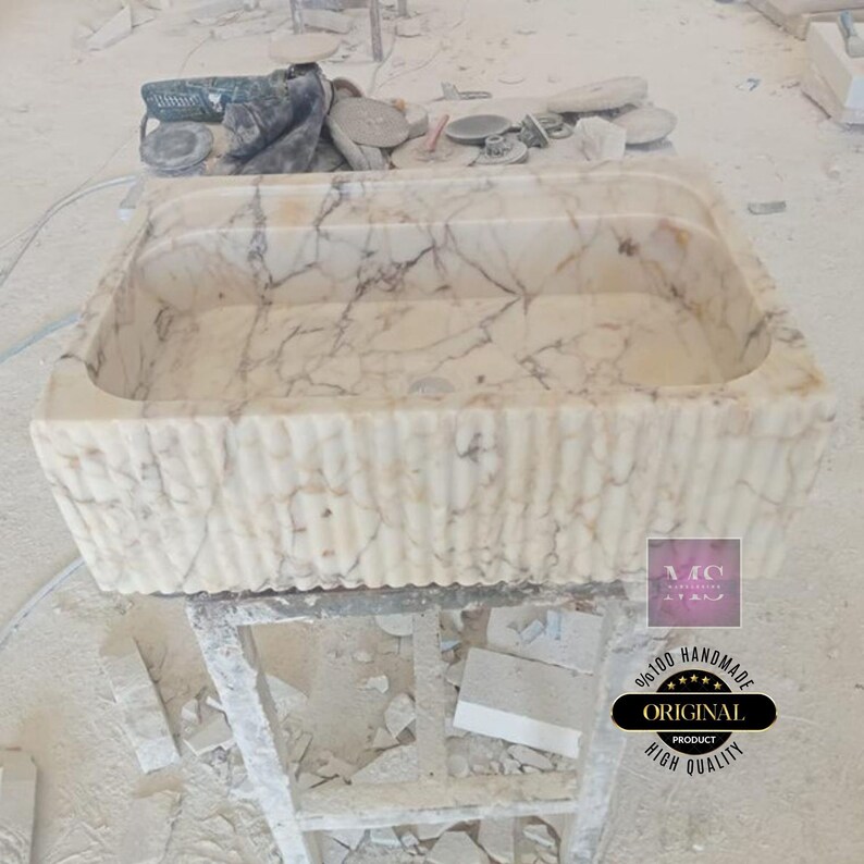 May include: A white marble sink with a rippled edge. The sink is handmade and has a high quality finish. The sink is sitting on a wooden stand.