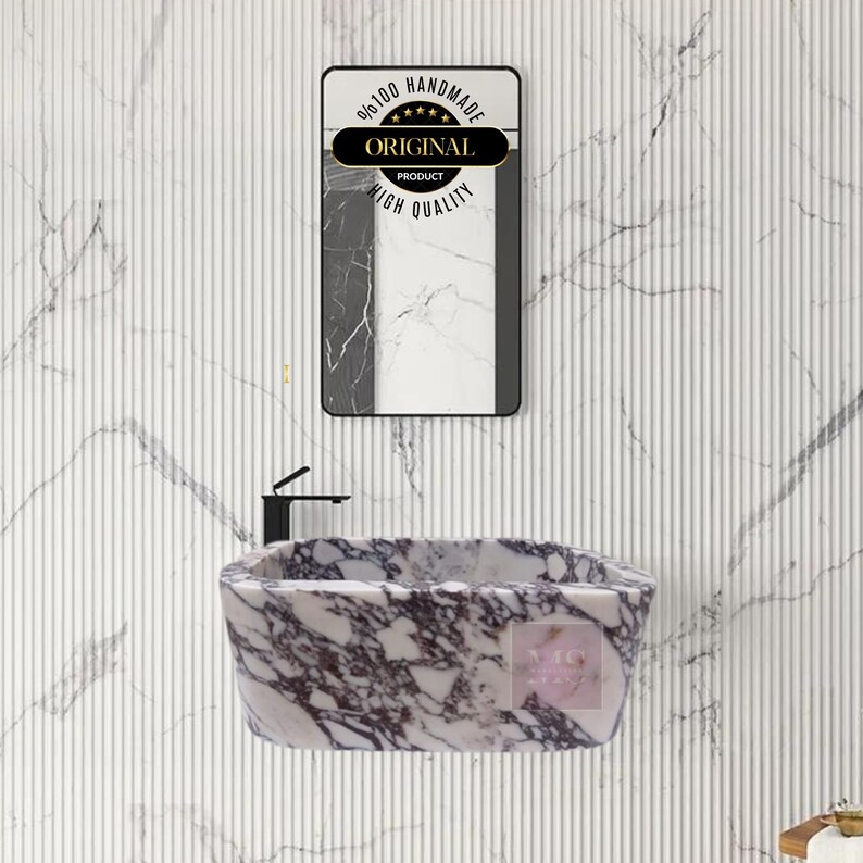 Op de afbeelding: Een witte marmeren wastafel met paarse en grijze aderen. De wastafel is ovaalvormig en heeft een gladde, gepolijste afwerking. De wastafel is aan een muur gemonteerd en heeft een zwarte kraan.
