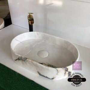 May include: White oval marble sink with a gold faucet and a drain stopper. The sink has a unique marbled pattern. The sink is labeled "Original Product High Quality Handmade".