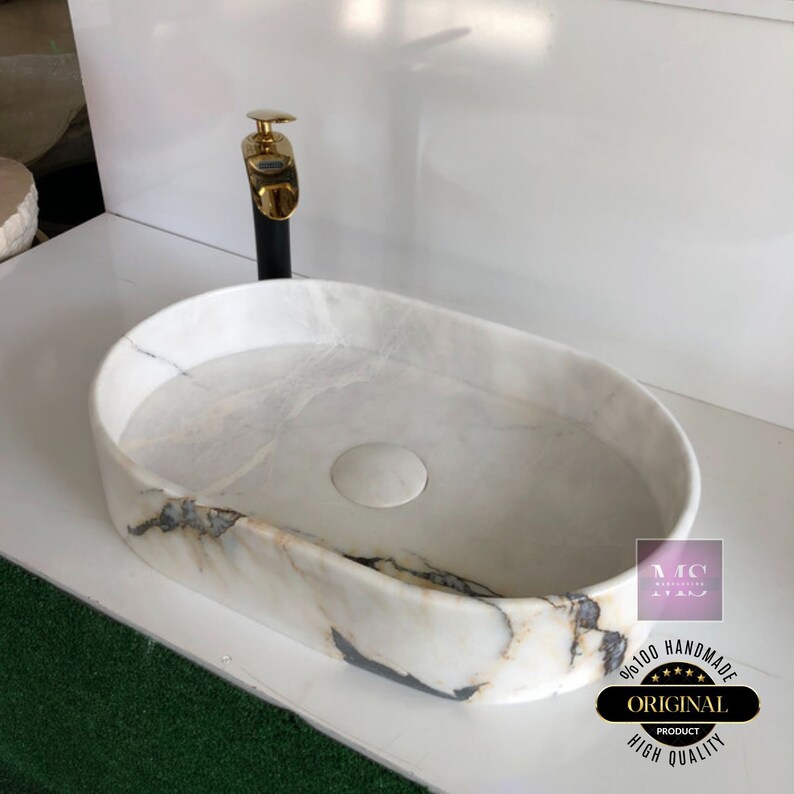 May include: White oval marble sink with a gold faucet and a drain stopper. The sink has a unique marbled pattern. The sink is labeled "Original Product High Quality Handmade".