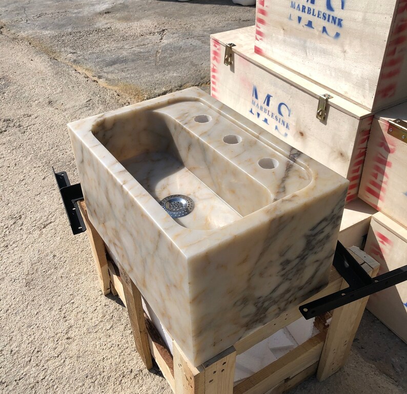Right three-quarter workshop view of a rectangular Calacatta Viola marble sink, showing the polished natural stone, deep basin, centered metal drain, three faucet holes and heavy-duty wall brackets.