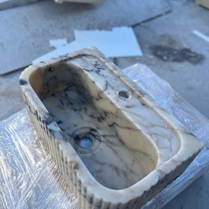 "Handmade Calacatta marble wall-mounted sink with fluted front design, shown in a minimalist bathroom setting. Featuring a single faucet hole and rear shelf for soap or accessories. Perfect for modern, luxury interiors. Custom sizes available. Polished interior, natural veining, and matte black faucet compatibility."