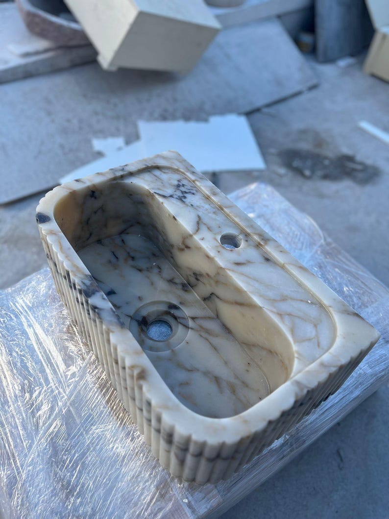 "Handmade Calacatta marble wall-mounted sink with fluted front design, shown in a minimalist bathroom setting. Featuring a single faucet hole and rear shelf for soap or accessories. Perfect for modern, luxury interiors. Custom sizes available. Polished interior, natural veining, and matte black faucet compatibility."