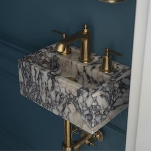 May include: A white marble vessel sink with a gold-toned faucet and drain. The sink is mounted on a wall and has a blue background.