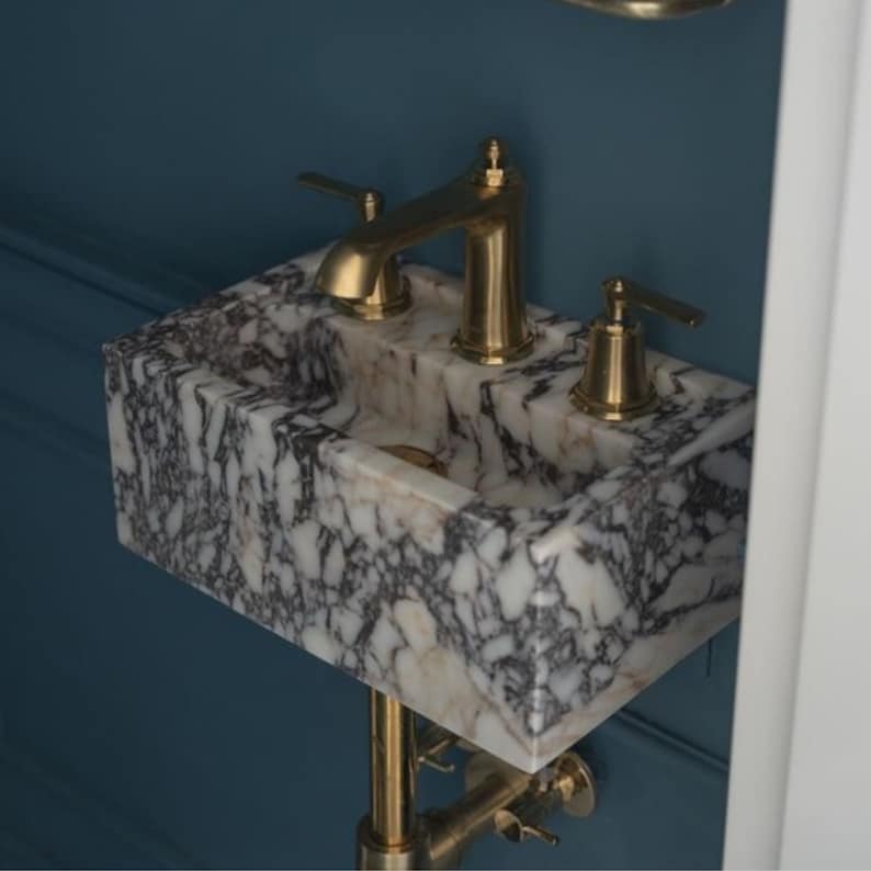 May include: A white marble vessel sink with a gold-toned faucet and drain. The sink is mounted on a wall and has a blue background.