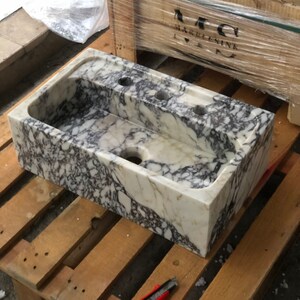Puede incluir: Un lavabo de m&aacute;rmol blanco con vetas moradas y doradas. El lavabo tiene cuatro agujeros para grifos y un agujero de desag&uuml;e en la parte inferior.