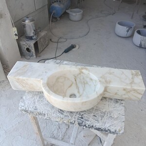 Luxurious wall-mounted Calacatta marble sink carved from a single block. Featuring 3 faucet holes, fluted front design, and floating vanity look. Ideal for modern, minimalist bathrooms. Custom sizes available. Handmade from natural Italian stone. Bathroom sink, marble vanity, stone basin, designer sink, fluted marble, wall mount sink, handmade sink, rectangular sink, luxury bathroom decor.