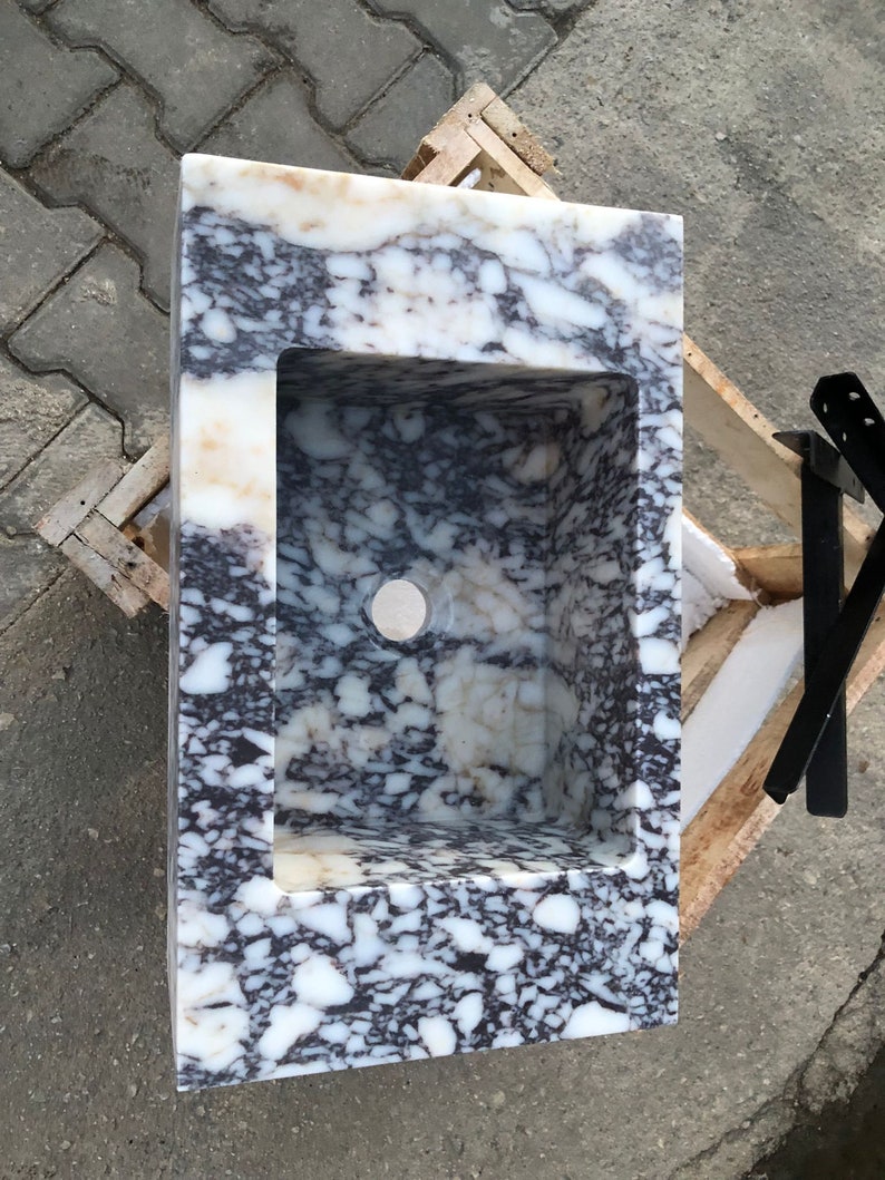 May include: A white marble sink with a black and gray marbling pattern. The sink has a rectangular shape and a rounded edge. The sink is set in a wooden frame.