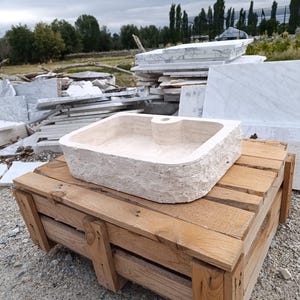 Op de afbeelding: Een rechthoekige beige stenen wastafel met een ruwe, natuurlijke buitenkant en een gladde, gepolijste binnenkant. De wastafel is voorzien van een enkel gecentreerd kraangat aan de achterrand en is vervaardigd uit lichtgekleurde natuursteen.