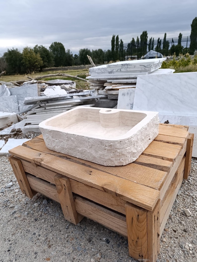 Op de afbeelding: Een rechthoekige beige stenen wastafel met een ruwe, natuurlijke buitenkant en een gladde, gepolijste binnenkant. De wastafel is voorzien van een enkel gecentreerd kraangat aan de achterrand en is vervaardigd uit lichtgekleurde natuursteen.