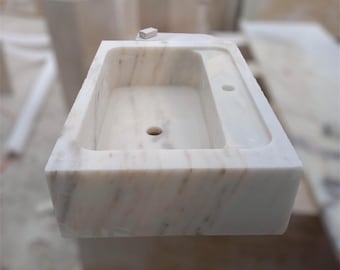 Puede incluir: Lavabo de baño de mármol blanco con forma rectangular y un solo agujero de drenaje. El lavabo tiene un borde elevado alrededor de la cuenca y un acabado liso y pulido.