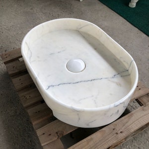 Może przedstawiać: Biała owalna umywalka marmurowa z białym ceramicznym korkiem odpływowym.