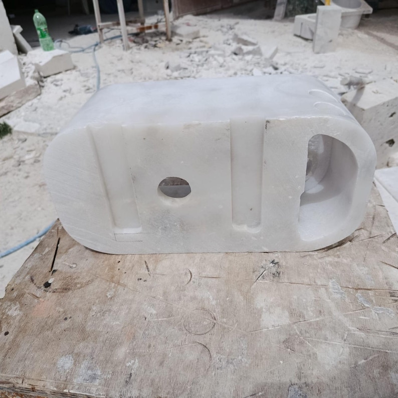 May include: A carved white marble sculpture with a rounded rectangular shape. The sculpture has a circular hole and a curved, hollowed-out section on one end. The piece is set on a wooden surface, suggesting a workshop or studio setting.