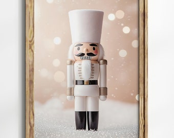 May include: A white wooden nutcracker with a gold belt and black boots stands on a sparkly white surface with a pink and white bokeh background. The nutcracker has a moustache and a red blush on its cheeks.