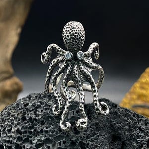 May include: A silver octopus ring with a textured head and tentacles. Two small, round, clear gemstones are set on the top of the ring. The ring is displayed on a dark, porous rock.