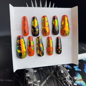 Pode incluir: Um conjunto de dez unhas postiças numa caixa branca. As unhas são uma mistura de laranja e preto, com detalhes em folha holográfica. Algumas unhas apresentam um design de rosto de abóbora, outras padrões de diamantes.
