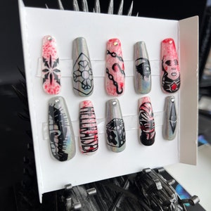 May include: A set of ten false nails with a holographic finish. Each nail is decorated with a different black and white graphic design, including a skull, a chain, a dice, and a snowflake. The nails are arranged in two rows on a white card.