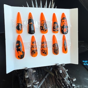 May include: Set of ten orange and black fake nails with various designs. The designs include a clock, a skull, a person with a mask, and the words "We Suggest You Show You".