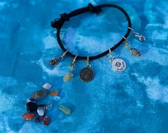 Könnte beinhalten: Ein schwarzes Kordelarmband mit mehreren Anhängern, darunter ein Anhänger mit dem Sternzeichen "Krebs", ein Anhänger mit der Aufschrift "69 Imagine" und ein Anhänger mit einem Spiralmuster. Außerdem liegen mehrere kleine, bunte Steine um das Armband herum.
