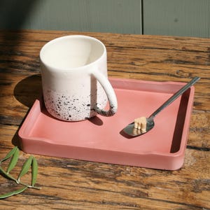 Peut inclure: Une tasse en céramique blanche avec des taches noires sur un plateau en béton rose avec une cuillère en argent et un morceau de sucre.
