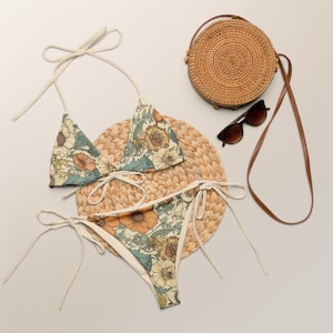 May include: A two-piece floral bikini set with a triangle top and tie-side bottoms. The bikini features a cream background with orange, yellow, and blue floral patterns. A round woven bag, sunglasses, and a woven placemat are also in the image.