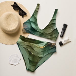 May include: A two-piece swimsuit with a green and gold abstract design. The top is a scoop-neck style, and the bottoms are high-waisted. The image also includes a straw hat, sunglasses, sunscreen, and a seashell, suggesting a beach or vacation theme.
