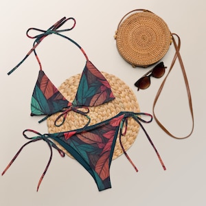 Op de afbeelding: Een tweedelige bikiniset met een bladerenpatroon in tinten teal, rood en bruin. De bikinitop is een driehoekmodel met strikbandjes en de broekjes zijn een klassieke snit met strikbandjes aan de zijkanten. Een ronde rieten tas en zonnebril zijn ook zichtbaar.