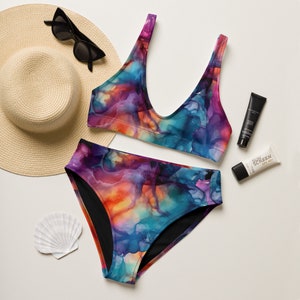 May include: A two-piece swimsuit with a vibrant abstract watercolor design in shades of purple, blue, orange, and pink. The swimsuit is paired with a straw hat, sunglasses, sunscreen, and a seashell, suggesting a beach or vacation theme.