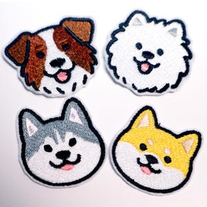 Op de afbeelding: Vier geborduurde hondenpatches. De patches tonen de gezichten van een bruin-witte hond, een witte pluizige hond, een grijs-witte husky en een geel-witte Shiba Inu. Elke patch heeft een zwarte omtrek en een glimlachende uitdrukking.