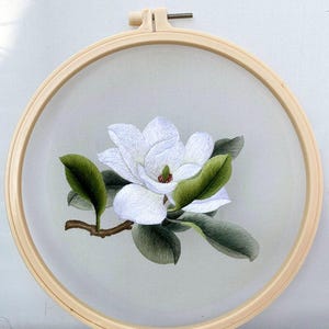 May include: An embroidered white magnolia flower with green leaves and a brown stem, displayed in a wooden embroidery hoop. The needlework highlights the delicate petals and the overall craftsmanship of the floral design.