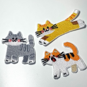 Può includere: Tre toppe ricamate a forma di gatto: un gatto grigio tigrato in piedi, un gatto giallo tigrato che si allunga con zampe bianche e un gatto bianco con strisce arancioni e nere e la bocca aperta. Ogni toppa ha baffi neri e guance rosa.