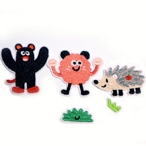Könnte beinhalten: Gestickte Aufnäher mit Cartoon-Tieren: ein schwarzer Bär mit roten Akzenten, ein rosa Wesen mit schwarzen Beinen und ein Igel mit grauen Stacheln. Ein grüner Busch und ein Blatt sind ebenfalls enthalten. Ideal zum Dekorieren von Kleidung.